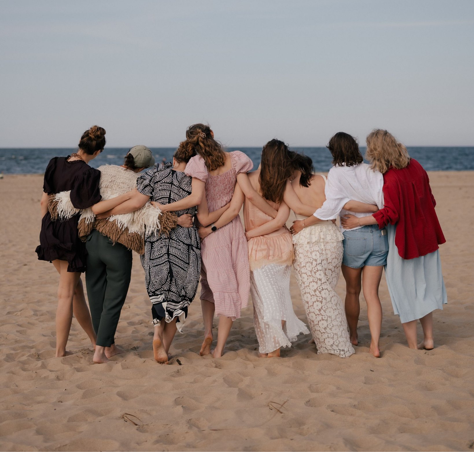 Grupa kobiet stoi objęta na plaży, tyłem do obiektywu, twarzą do morza.